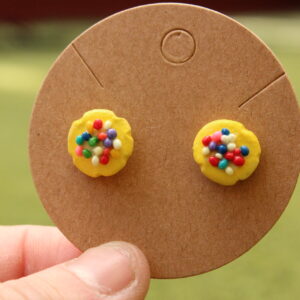 An example of the cookie earrings descirbed are shown. The cookies are yellow with rainbow sprinkles. They are affixed to a cardboard display card.