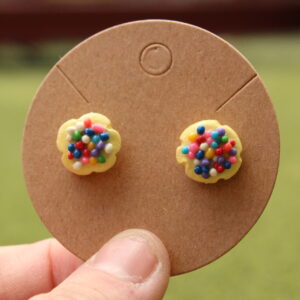 An example of the cream sugar cookie earrings is shown. The earrings are displayed on a cardboard card, and each feature a tiny cream colored sugar cookie topped with rainbow sprinkles.